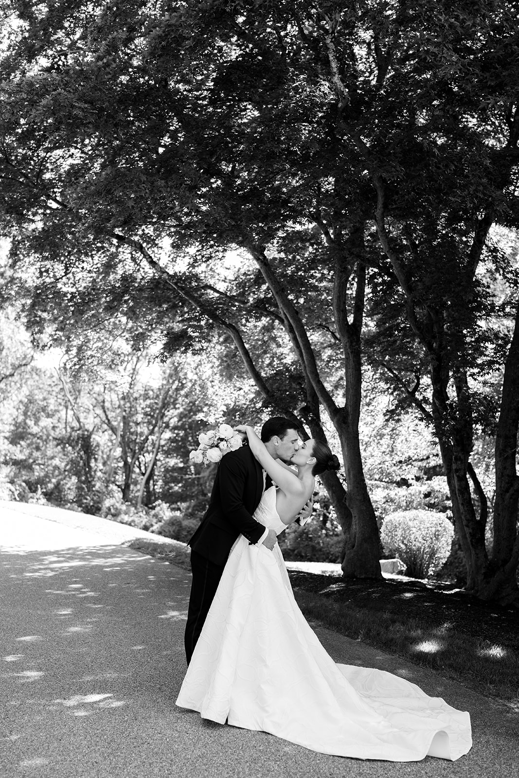Black and white film photo of the groom dipping and kissing the bride on a shaded pathway.